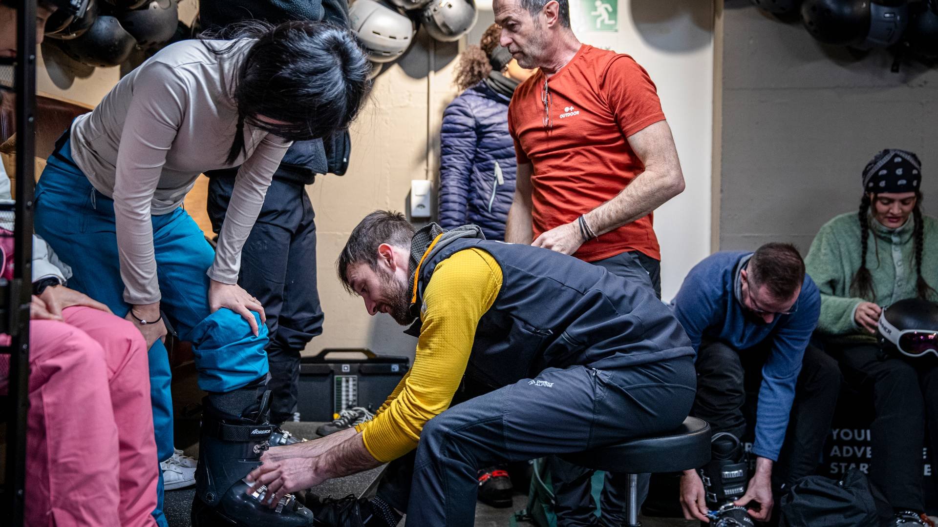 People fitting ski boots indoors, surrounded by helmets and winter gear, preparing for an activity.